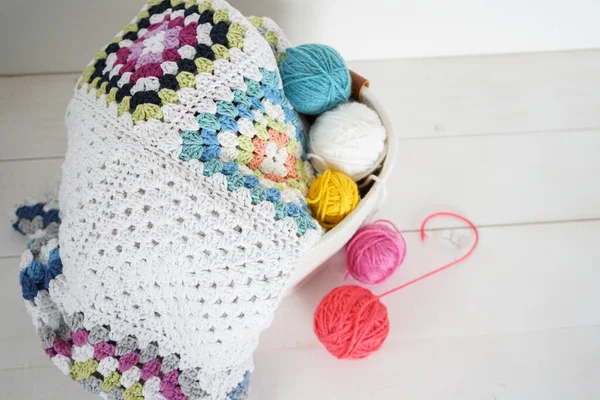 Beyaz ahşap zeminde yün topları olan beyaz sepetli büyük anne battaniyesi.