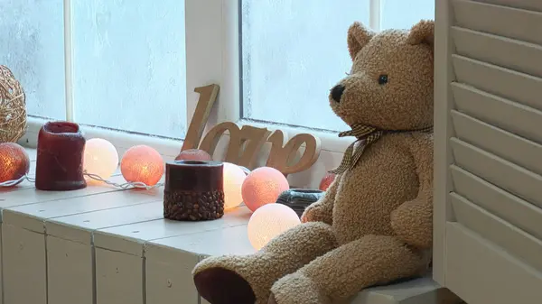 A teddy bear is sitting by a frozen window. Next to the bear, the word love is carved out of plywood. There is a garland on the windowsill