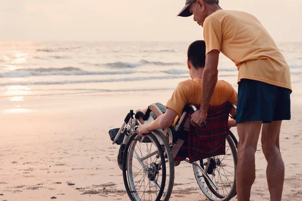 A parent, caregiver or volunteer helping teenage boy push wheelchair to the beach in evening sunset,exercise and learning about nature on the sea,Travel on vacation with family, Mental health concept.