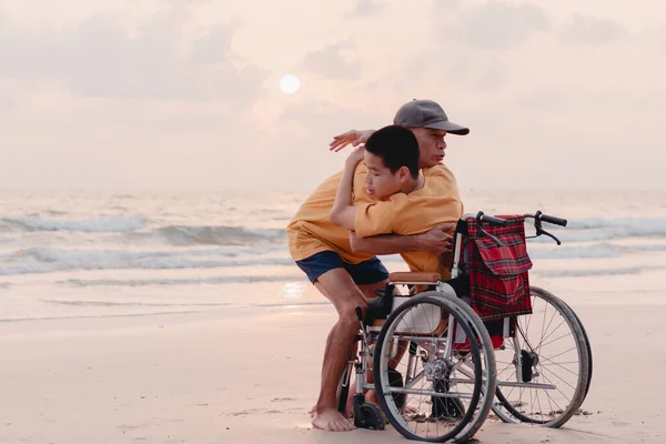 A parent, caregiver or volunteer help moving boy to standing from wheelchair on sunset sea beach,Beautiful moment,sharing,ministration,caring,Travel on vacation with family, Good mental health concept
