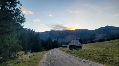 Kırsal bir köy yolu ve Polonya 'nın Zakopane yakınlarındaki Tatra Dağları' ndaki bir dağ vadisinde gün batımında geleneksel ahşap çoban kulübeleri..