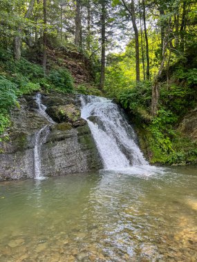 Sahne görkemli bir şelaleyi ihtişamlı bir şekilde yakalar, sanki zamanda donmuşçasına. Kristal berrak su, kayalık bir uçurumdan düşer. Köpüklü beyaz püskürtüsü, etrafındaki arazinin toprak tonlarına zıt düşer..