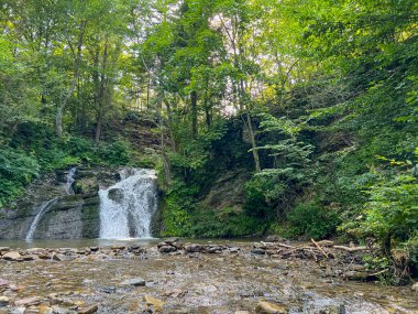 Sahne görkemli bir şelaleyi ihtişamlı bir şekilde yakalar, sanki zamanda donmuşçasına. Kristal berrak su, kayalık bir uçurumdan düşer. Köpüklü beyaz püskürtüsü, etrafındaki arazinin toprak tonlarına zıt düşer..