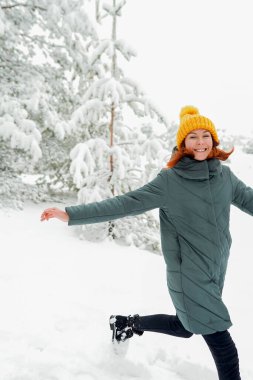 Sıcak elbiseli, kızıl saçlı neşeli kız dışarıda karla oynuyor. Güzel ormanın yakınında.