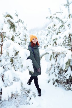 Sıcak elbiseli, kızıl saçlı neşeli kız dışarıda karla oynuyor. Güzel ormanın yakınında.