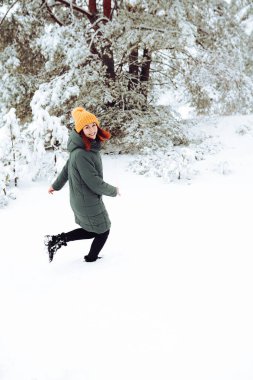 Sıcak elbiseli, kızıl saçlı neşeli kız dışarıda karla oynuyor. Güzel ormanın yakınında.