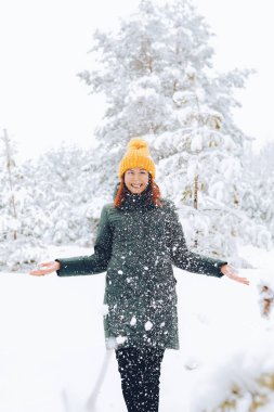 Sıcak elbiseli, kızıl saçlı neşeli kız dışarıda karla oynuyor. Güzel ormanın yakınında.