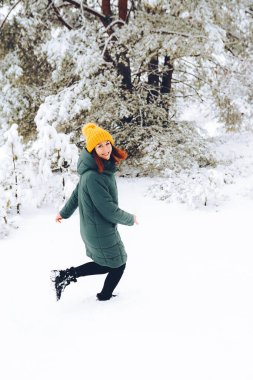 Sıcak elbiseli, kızıl saçlı neşeli kız dışarıda karla oynuyor. Güzel ormanın yakınında.