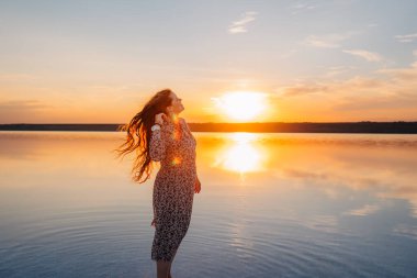 Uzun, rüzgarlı saçları olan genç bir kadın sakin bir gölün kenarında huzur içinde gülümsüyor altın bir günbatımında, huzurlu bir atmosferde özgürlük, mutluluk ve doğal güzellik yayıyor..