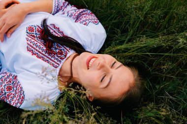 Genç bir kadın yemyeşil bir tarlada dinlenir, nakışlı bir bluz giyer, neşe ve sükunet yayar ve huzurlu bir atmosferin tadını çıkarır..