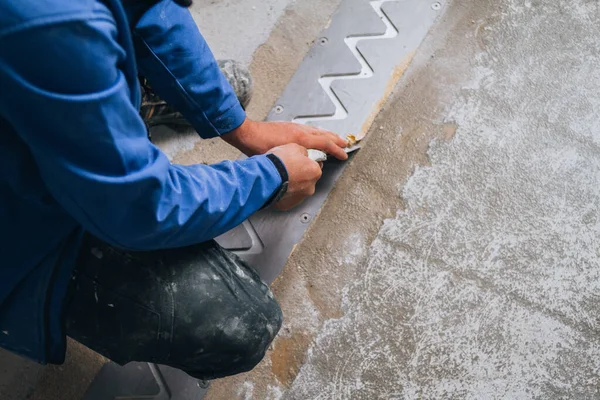 worker\'s skilled hands installing floor expansion joints. The worker is shown wearing protective gear and kneeling down on the concrete floor, using specialized tools to carefully measure and cut the material to fit precisely.