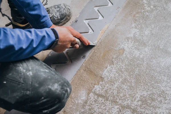 worker\'s skilled hands installing floor expansion joints. The worker is shown wearing protective gear and kneeling down on the concrete floor, using specialized tools to carefully measure and cut the material to fit precisely.