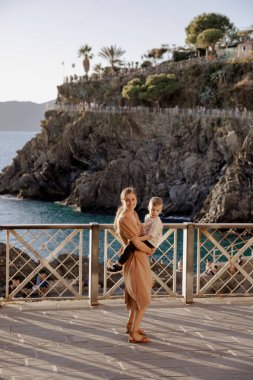 Anne, İtalya 'nın büyüleyici sahili olan 5 Terre' de tatildeyken küçük oğlunu kollarında tutuyor. Fotoğraf, Ligurian Denizi 'nin nefes kesici manzarasını ve köylerin renkli binalarını yakalıyor.