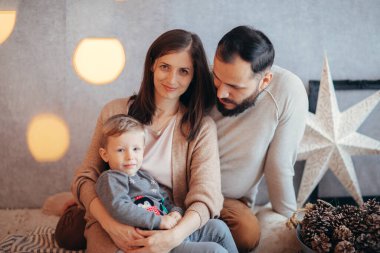 Bu yılki Noel için neşeli bir şekilde süslenmiş evlerinde, ışıl ışıl genç bir aile, bir dizi hediye ve parıldayan ışıklar arasında 2 yaşındaki sarışın oğlanlarını kucaklıyor, neşe ve beraberliğin özünü yakalıyor.