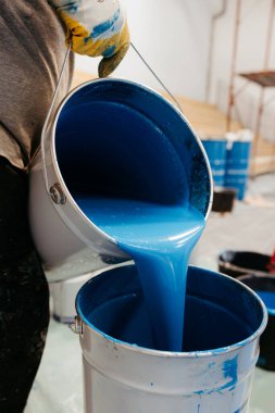 Construction worker is pouring blue epoxy resin from a bucket into another bucket to prepare the mixture for flooring clipart