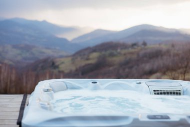 Bulutlu bir günde vadiye ve dağlara bakan ahşap bir güvertede modern jakuzi fokurduyor.