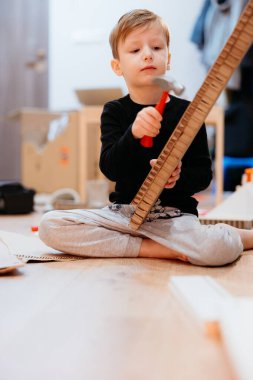 Kartonla oyuncak ev yapmaya odaklanmış, oyun odasının zemininde oturan genç bir çocuk.