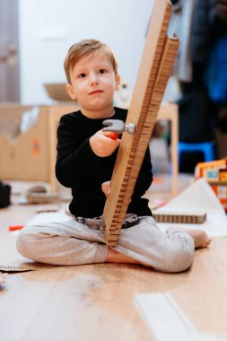 Genç bir çocuk ahşap inşaatı topluyor. Konsantrasyonuyla birlikte bir oyun odasında yerde oturuyor.