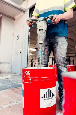 İnşaat işçisi, inşaat alanında matkapla kırmızı kovaya epoksi reçinesi karıştırıyor.