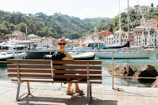 Turist bir bankta oturmuş Portofino, İtalya limanındaki renkli binaların ve teknelerin manzarasının keyfini çıkarıyor.