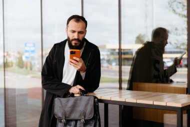 Sakallı işadamı, evrak çantasını tutarken ve modern ofis binasının dışındaki masaya yaslanırken akıllı telefondan mesajlar okuyor.