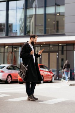 Modern binaların önünden geçerken akıllı telefon kullanan omuz çantalı sakallı bir işadamı.