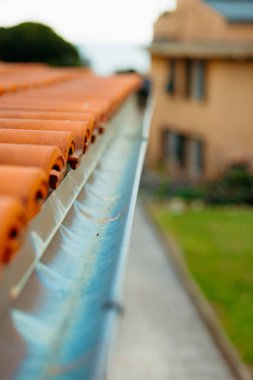 Metal yağmur oluğu kil çatı kiremitlerinden akan suyu topluyor. Arka planda bulanık bir ev ve yeşil çimen var.