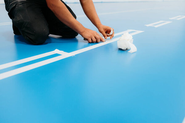 Строитель, маркирующий белые линии на синем эпоксидном покрытии пола с помощью маскировочной ленты и резака
