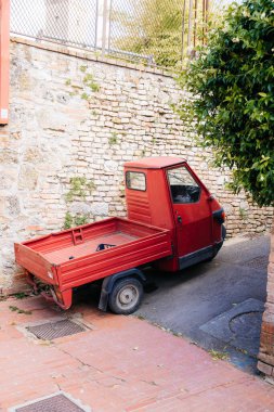 İtalya 'da eski bir Toskana şehrinin dar bir sokağında, tuğla bir duvarın yanına park edilmiş küçük kırmızı üç tekerlekli bir kamyon.