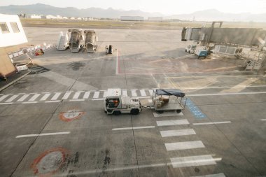 Havaalanı pistinde yük vagonunu çeken çekici. Yolcu köprüleri ve arka planda dağlar var.