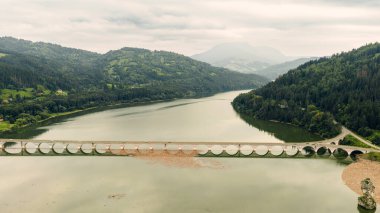 Yeşil tepeler ve bulutlu gökyüzüyle çevrili sakin sulara yansıyan uzun bir göl.