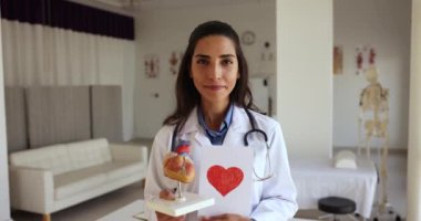Neşeli genç Latin kardiyolog elinde kırmızı kalp ve vücut organı modeli olan bir kağıt tutarak dişlek gülümsemeyle kameraya bakıyor, doktorların gününü kutluyor, kalp muayenesini destekliyor.