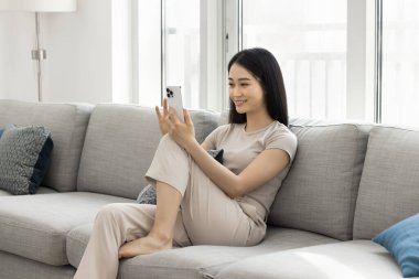 Mutlu, genç, Asyalı blog yazarı kadın kendini videoya kaydediyor, selfie çekiyor, akıllı telefondan konferans görüşmesi yapıyor, cep telefonunu kanepe üzerinde kullanıyor, diz destekli bir cihaz tutuyor.