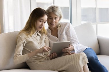 Anne ve genç kız hafta sonları dijital tablet kullanıyor, rahat bir kanepede birlikte oturuyor, web sörfü yapıyor, hizmetler veya hediyeler alıyor, yeni başvurular alıyor, internette boş vakitlerini evde geçiriyorlar.