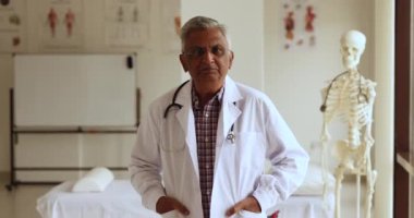 Ciddi derecede kıdemli Hintli doktor beyaz üniformalı adam hastane ofisinde duruyor, ellerini ceplerinde tutuyor, mutlu oluyor, pozitif oluyor, kameraya bakıyor, gülümsüyor, profesyonel portre için poz veriyor.