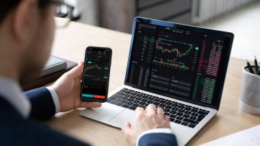 Erkek tüccar, bilgisayar grafiği ve grafiği kullanarak, yatırımı analiz ederek, borsa bilgilerini kontrol ederek, borsa verilerini içeren akıllı bir telefon başında oturuyor. Büyüme, kâr, uygulamalar