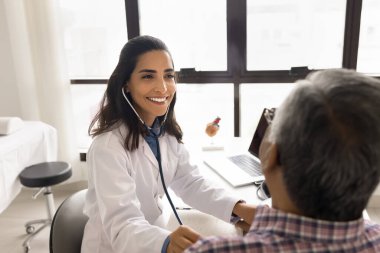 Pozitif güzel genç pratisyen kadın kıdemli hastayı muayene ediyor, steteskop takıyor, kalp atışlarını, nefes alış verişini, akciğerlerini kontrol ediyor, çalışma masasında oturuyor.