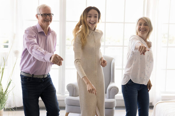 Happy pre-teen girl dancing with mum 70s grandfather in living room, listen energetic disco music, actively move to song, enjoy favorite hobby. Overjoyed three different ages people having fun at home