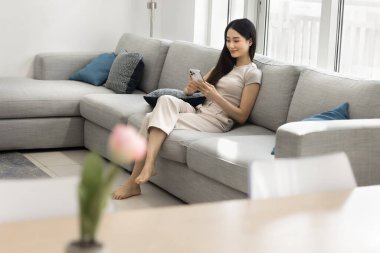 Pozitif genç Koreli kadın cep telefonuyla konuşuyor, evdeki büyük rahat koltukta dinleniyor, modern teknolojinin tadını çıkarıyor, cep telefonunda akıllı ev uygulaması kullanıyor.