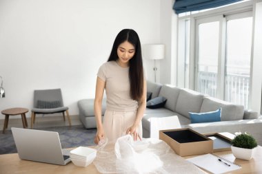 Olumlu Asyalı genç müşteri paketini evde açıyor, internet mağazasından paketleri açıyor, plastik ambalajı tutuyor, kırılgan tabakları kağıt kutuya koyuyor, iş yerinde dizüstü bilgisayarla duruyor.