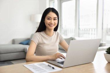 Mutlu Asyalı kız öğrenci evde dizüstü bilgisayar okuyor, online eğitim için elkazanç platformunu kullanıyor, bilgisayarla masaya oturuyor, kameraya bakıyor, daktilo ediyor, gülümsüyor.