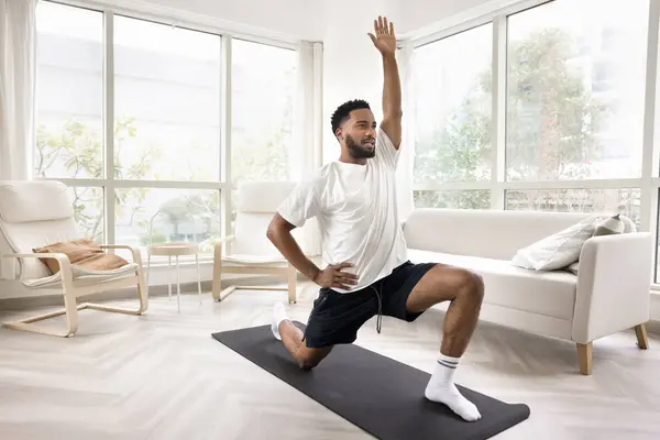 Positive motivated athletic young African man enjoying fitness activity on yoga mat at home, doing lunge, exercises, training body, keeping fit, stretching legs, working on hips flexibility