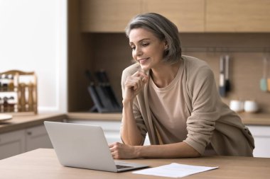 Gülümseyen güzel kadın mutfak masasında dizüstü bilgisayara bakıyor, e-posta ile haber veya ticari teklif okuyor, düşünce stratejisi yapıyor, serbest iş yapıyor, uzaktan sohbet ediyor, online proje üzerinde düşünüyor.