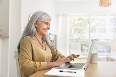 Neşeli, yaşlı, İspanyol, serbest çalışan bir iş kadını uzaktan işten zevk alıyor, dizüstü bilgisayarda yazıyor, gülümsüyor, gülüyor, serbest bir proje üzerinde çalışıyor, internetten eğitim alıyor.