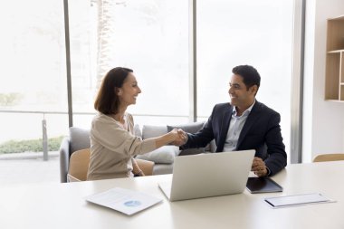 Mutlu başarılı iş ortakları iş yerinde el sıkışıyor, laptopta buluşma konusunda anlaşmaya varıyor. Olgun ve genç meslektaşlar işbirliği, ortaklık, gülme konularını tartışıyor