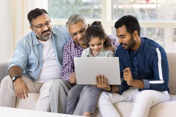 Cheerful Indian little kid girl, young dad, senior granddad, old grandpa using online application on laptop together, sitting close on sofa with computer, discussing Internet communication