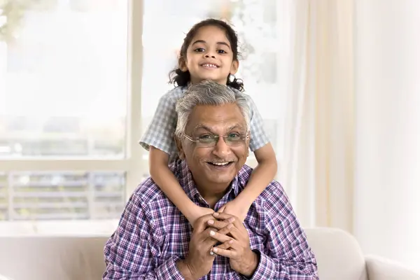 Cheerful pretty little granddaughter girl hugging grandpa from behind. Happy Indian grandfather piggybacking kid, looking at camera, smiling, enjoying leisure with child, posing for family portrait