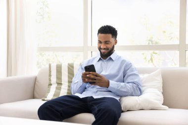 Pozitif Afrikalı genç bir adam cep telefonuyla yazıyor, koltukta dinleniyor, evdeki internet teknolojisini kullanıyor, online finans servisi, internet iletişimi uygulaması, sohbet ediyor, gülümsüyor, gülüyor.