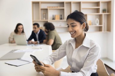 Kendine güveni tam profesyonel bir kız. Ofis iş arkadaşı masasında akıllı telefon kullanıyor. Toplantı odasında çeşitli iş arkadaşlarıyla oturup arka planda konuşuyor.