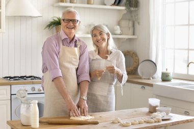 Önlüklü, orta yaşlı güzel bir kadın, olgun yaşlı bir babayla samimi, modern bir mutfakta yemek pişiriyor, kameraya bakıp gülümsüyor, birlikte eğleniyor, evde tatlı hazırlıyor. Mutfak, kravat, aile tatili.